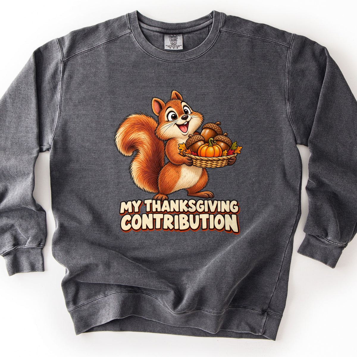 My Thanksgiving Contribution Comfort Colors crewneck sweatshirt in Pepper, laid flat. A smiling cartoon squirrel holds a woven basket of acorns, a small pumpkin, and autumn leaves above the words My Thanksgiving Contribution in cream letters. Shown on a plain white background.