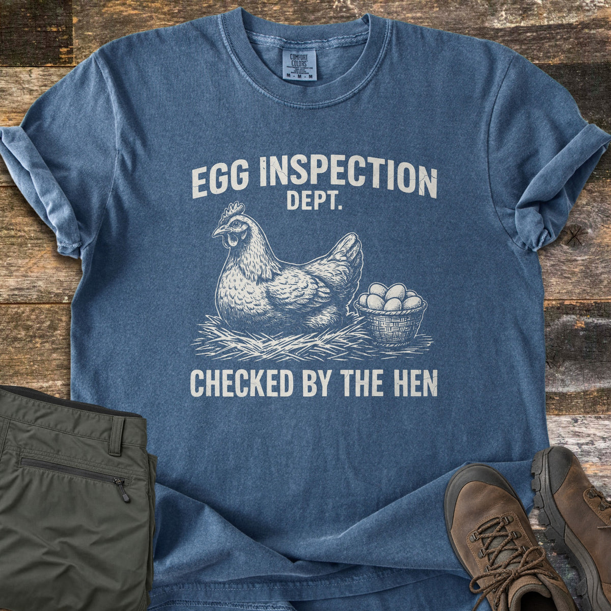 Egg Inspection Dept Comfort Colors T-shirt in Denim, laid flat. A sketched hen sits on straw beside a basket of eggs, between the words Egg Inspection Dept. and Checked by the Hen. Olive cargo pants and brown hiking boots lie beside it on weathered wood planks.