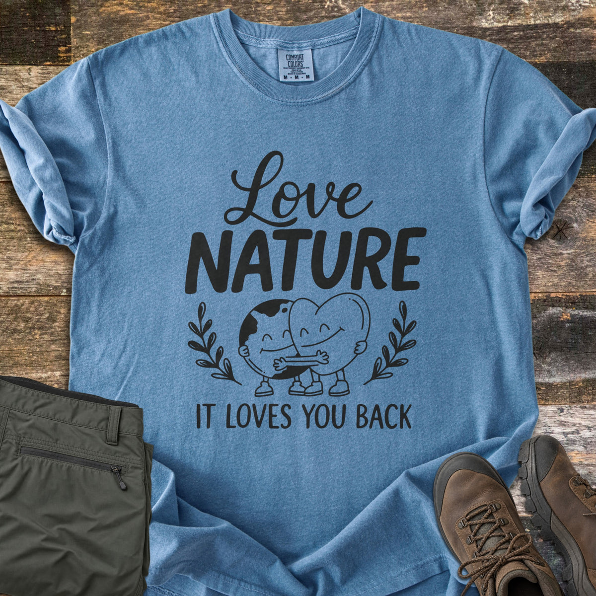 Nature Loves You Back Comfort Colors T-shirt in Blue Jean, laid flat. Black print reads Love Nature above a smiling cartoon Earth and heart hugging between leafy sprigs, with It Loves You Back below. It lies on weathered wood beside olive hiking pants and brown boots.