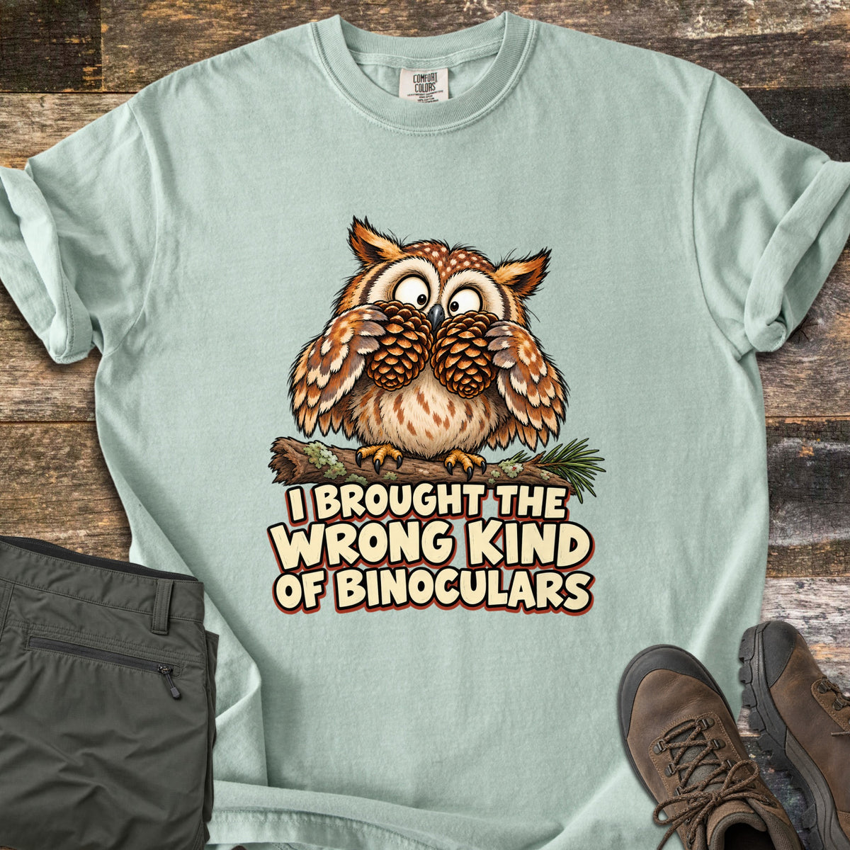 I Brought The Wrong Kind Of Binoculars Comfort Colors T-shirt in Bay, laid flat. A wide-eyed cartoon owl on a mossy branch holds two pinecones to its eyes like binoculars above the words I Brought The Wrong Kind Of Binoculars. Olive hiking pants and brown hiking boots lie beside it on weathered wood.