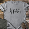 Bird Hunter Comfort Colors T-shirt in Grey, laid flat. A black heartbeat line crosses the chest with a hunter silhouette in a cap holding a shotgun and birds flying up from the line. Folded olive pants and brown hiking boots lie beside it on weathered wood.