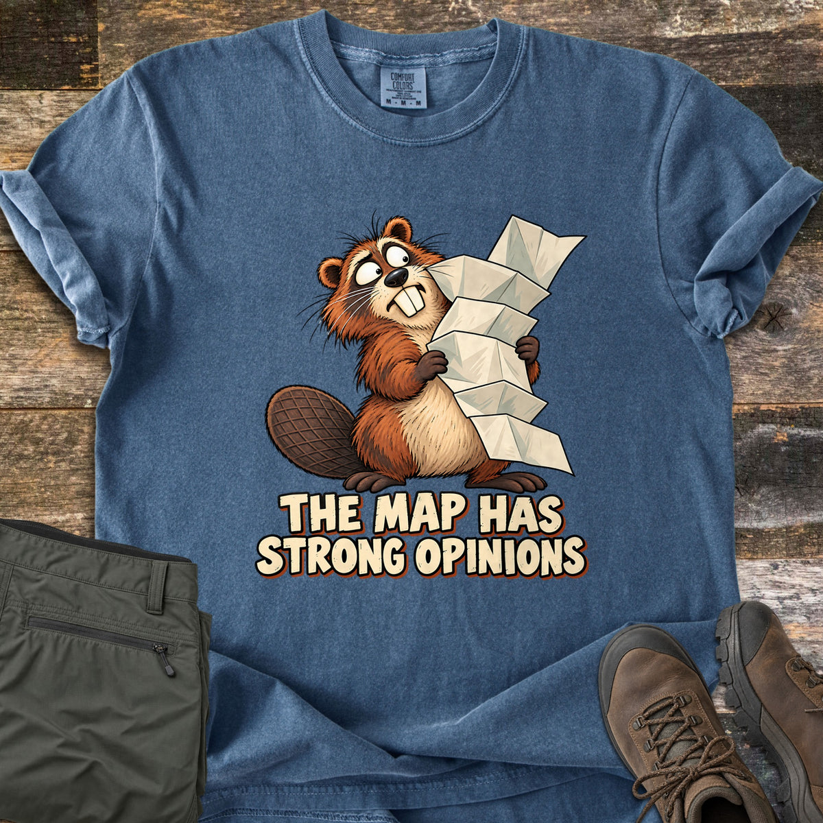 The Map Has Strong Opinions Comfort Colors T-shirt in Denim, laid flat. A wide-eyed cartoon beaver clutches a tall, unfolding paper map above the words The Map Has Strong Opinions. Olive hiking pants and brown hiking boots sit beside it on weathered wood planks.