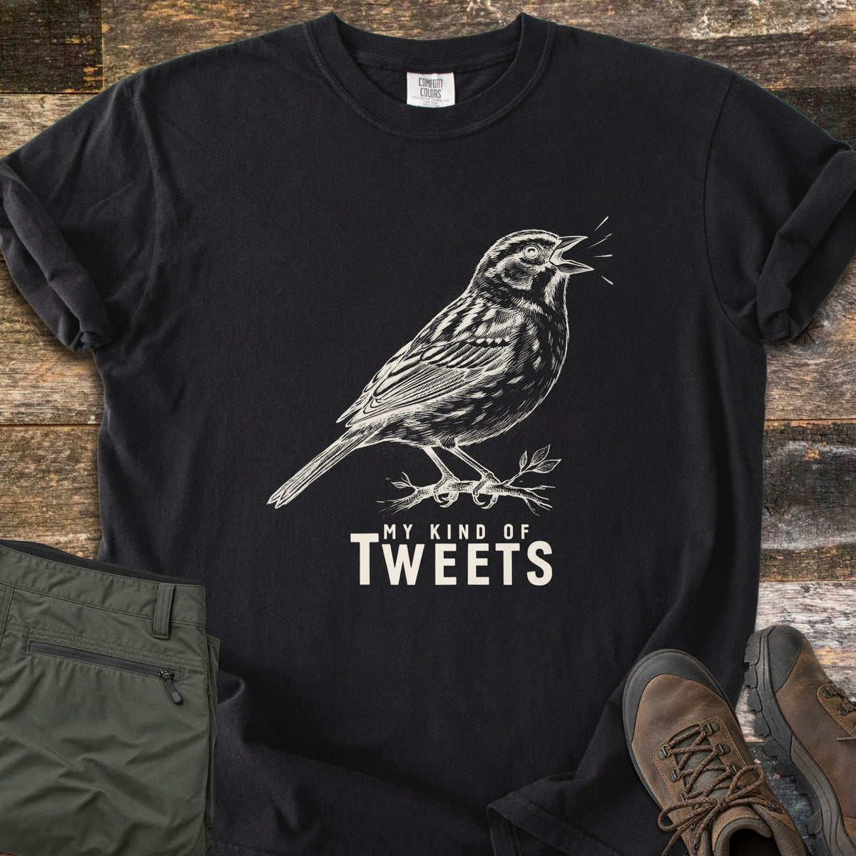 My Kind Of Tweets Comfort Colors T-shirt in Black, laid flat. A cream sketch of a singing sparrow perched on a leafy twig, above the words My Kind Of Tweets. Shown on rustic wood with folded olive hiking pants and brown hiking boots.