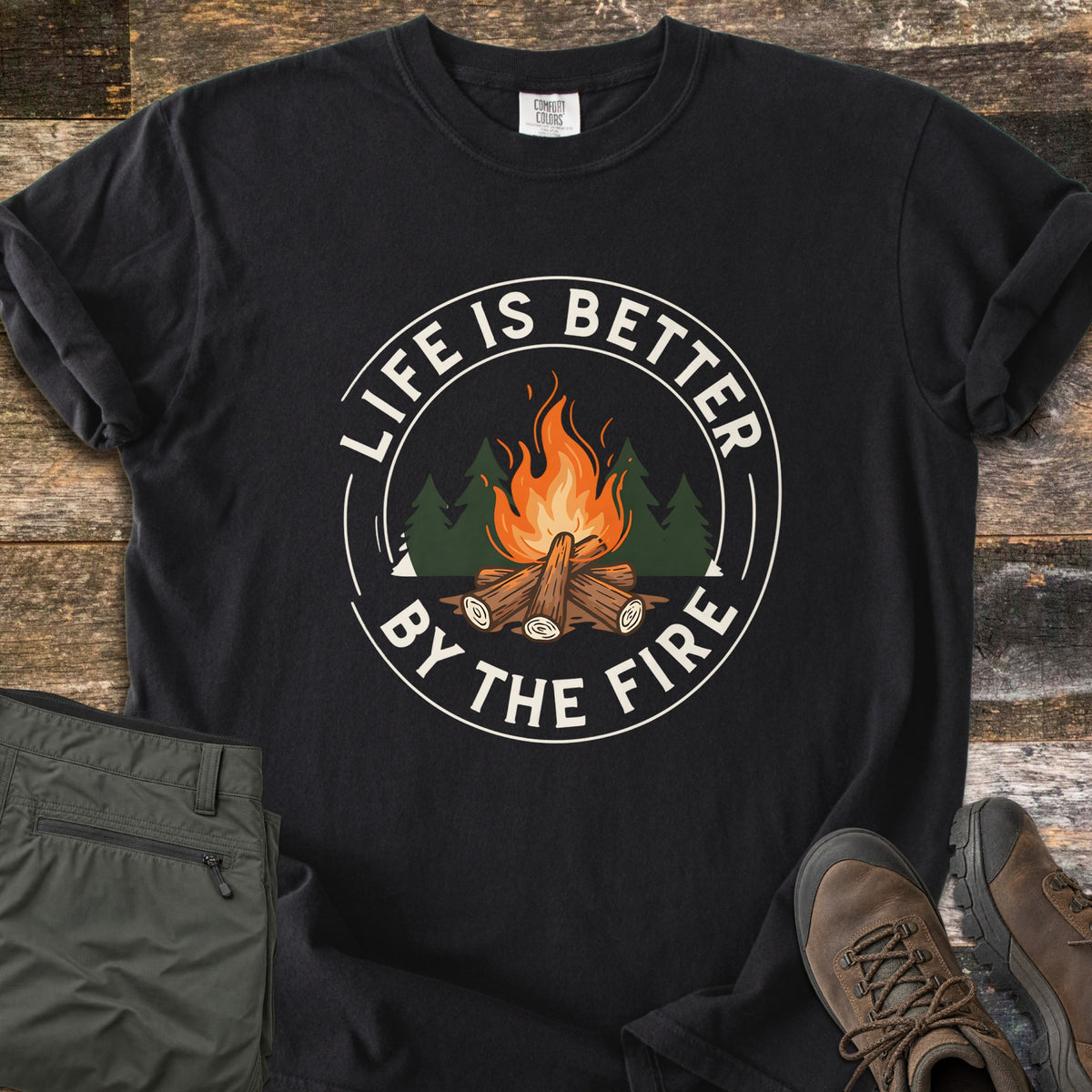 Life Is Better By The Fire Ring Comfort Colors T-shirt in Black, laid flat. A round badge shows an orange log campfire in front of dark green pine trees, ringed in white by the words Life Is Better By The Fire. Olive hiking pants and brown hiking boots lie beside it on weathered wood planks.
