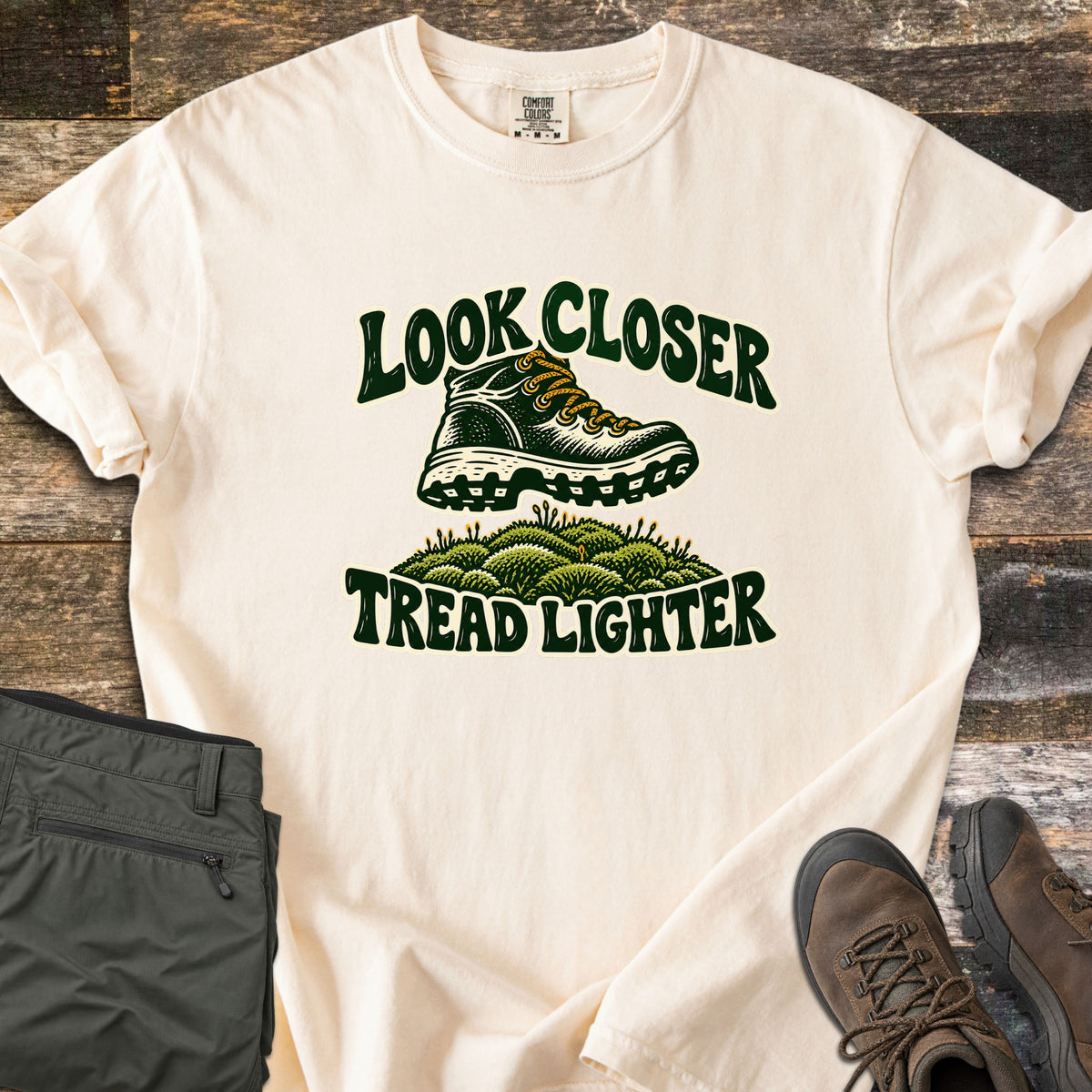 Look Closer Tread Lighter Comfort Colors T-shirt in Ivory, laid flat. A green hiking boot with yellow laces steps above a mound of moss between the words Look Closer and Tread Lighter. Sleeves rolled, on weathered wood with folded olive hiking pants and brown boots.