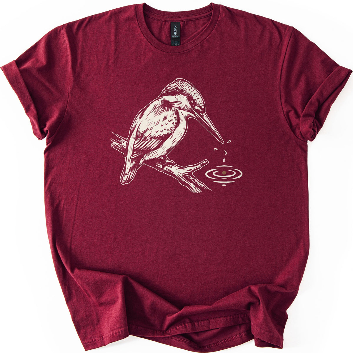 Kingfisher Comfort Colors T-shirt in Maroon, laid flat. A cream-colored drawing of a kingfisher perched on a branch, with droplets falling from its beak into rippling water below. The sleeves are cuffed and the shirt lies on a white background.