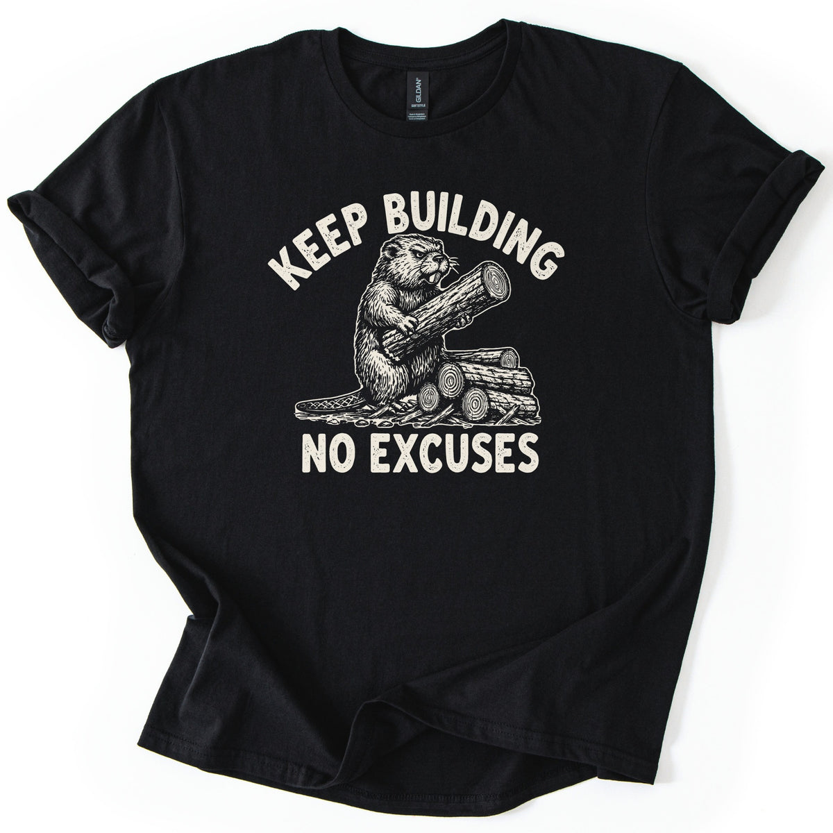 Keep Building Beaver Comfort Colors T-shirt in Black, laid flat. Cream sketch-style artwork of a beaver hugging a log while sitting on a pile of cut logs, with Keep Building arched above and No Excuses below. Sleeves are rolled on a white background.