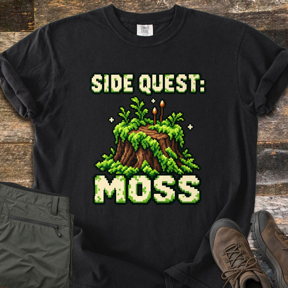 Side Quest Moss Comfort Colors T-shirt in Black, laid flat. Pixel art of a moss-covered tree stump with ferns, two small sporophytes and sparkles, between the words Side Quest: and Moss. Olive cargo pants and brown hiking boots lie beside it on weathered wood planks.