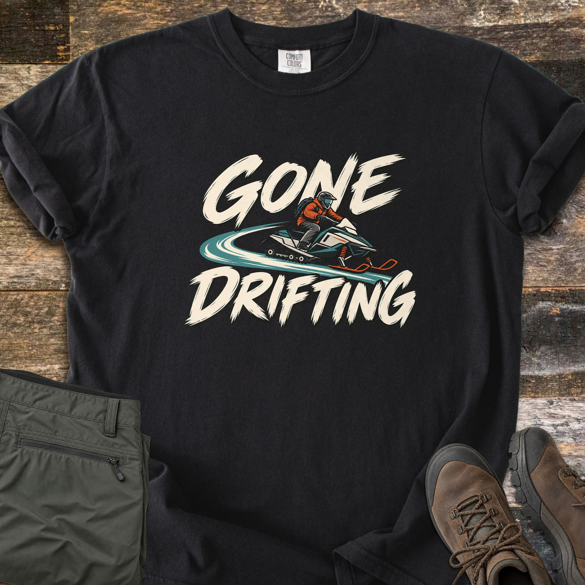 Gone Drifting Comfort Colors T-shirt in Black, laid flat. Cream brush letters read Gone Drifting around a helmeted rider in an orange jacket on a teal snowmobile with a swirling snow trail. Sleeves rolled, on weathered wood with folded olive pants and brown hiking boots.