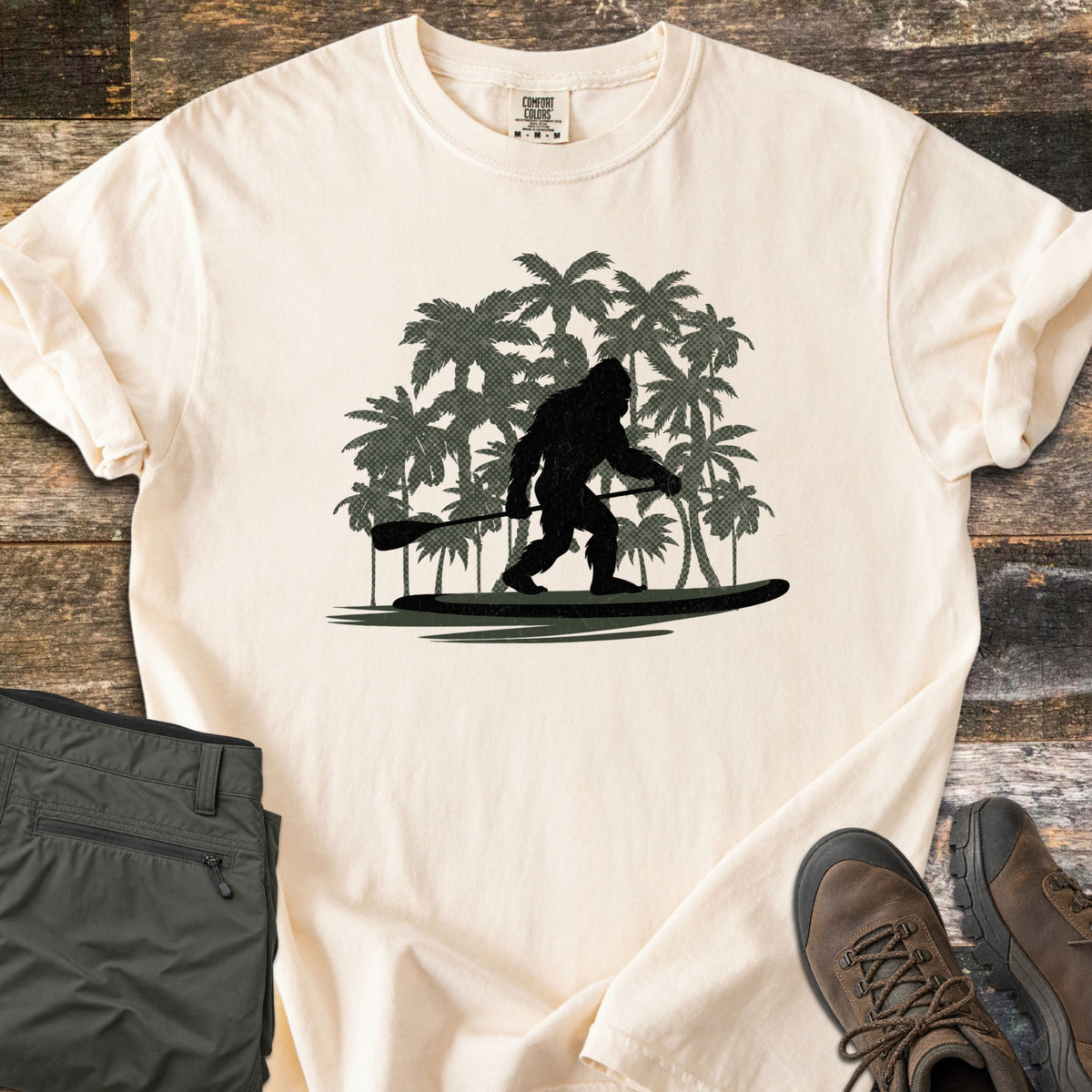 Paddle Board Squatch Comfort Colors T-shirt in Ivory, laid flat. A black silhouette of Bigfoot standing on a paddle board and holding a paddle, in front of dark green palm trees. Olive pants and brown hiking boots lie beside the shirt on weathered wood planks.