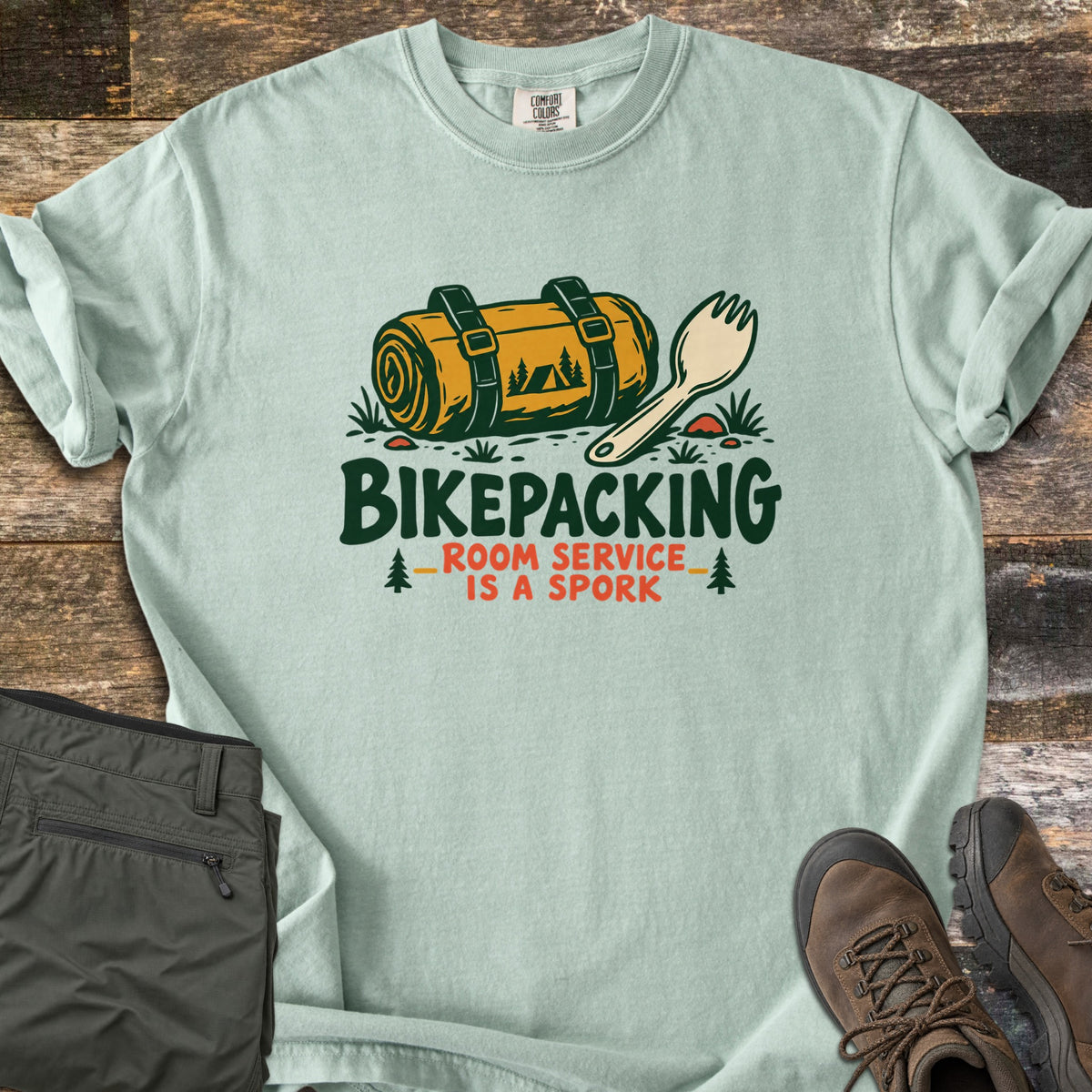 Bikepacking Room Service Is a Spork Comfort Colors T-shirt in Bay, laid flat. A yellow bedroll in dark green straps and a cream spork rest on grass above the words Bikepacking Room Service Is a Spork, flanked by small pines. Olive pants and brown hiking boots sit on weathered wood.