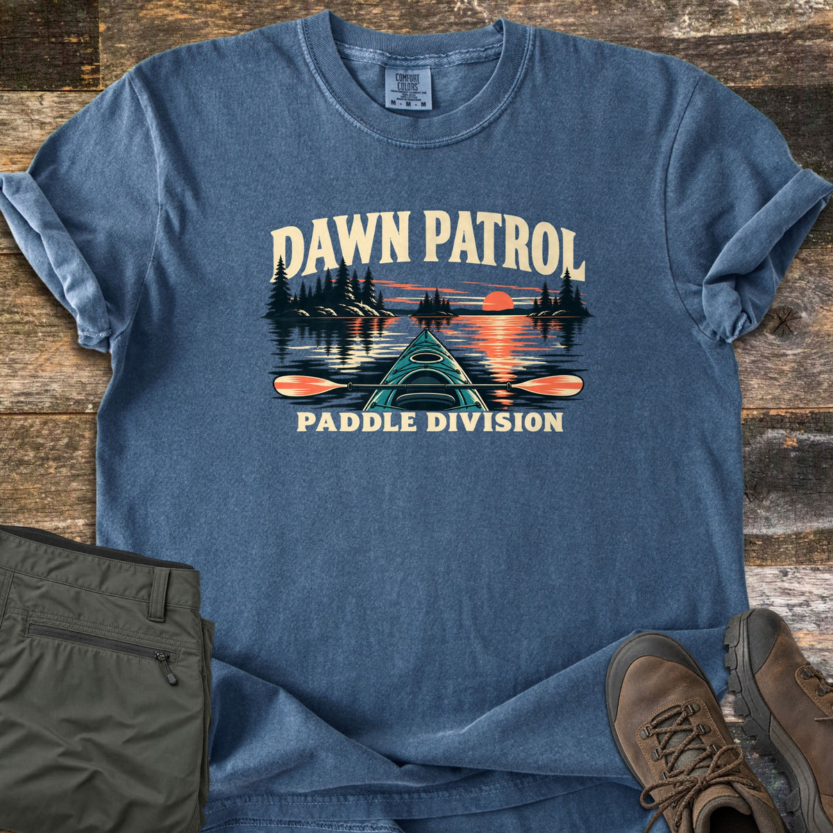 Dawn Patrol Paddle Division Comfort Colors T-shirt in Denim, laid flat. The words Dawn Patrol arch over a teal kayak bow and paddle on a lake with pine islands and an orange sunrise, above the words Paddle Division. Olive hiking pants and brown hiking boots lie beside it on weathered wood.