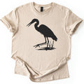 Great Blue Heron Comfort Colors T-shirt in Sand, laid flat. A black silhouette of a heron wading in shallow water beside cattails, beak open and one foot reaching forward. The shirt lies on a white surface with cuffed sleeves.