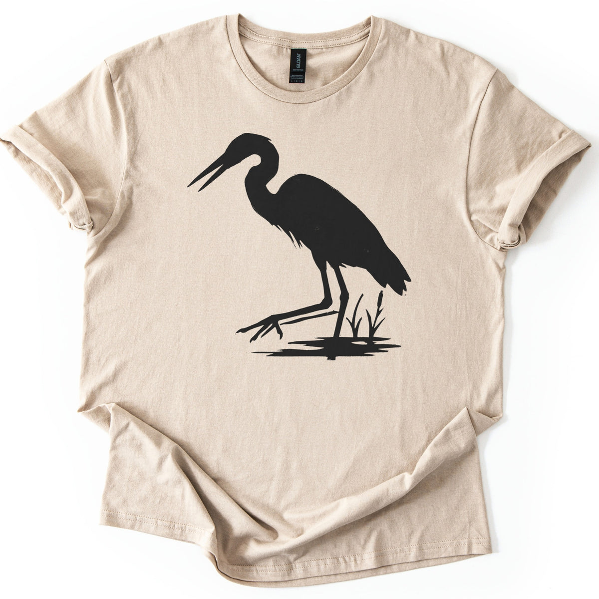 Great Blue Heron Comfort Colors T-shirt in Sand, laid flat. A black silhouette of a heron wading in shallow water beside cattails, beak open and one foot reaching forward. The shirt lies on a white surface with cuffed sleeves.