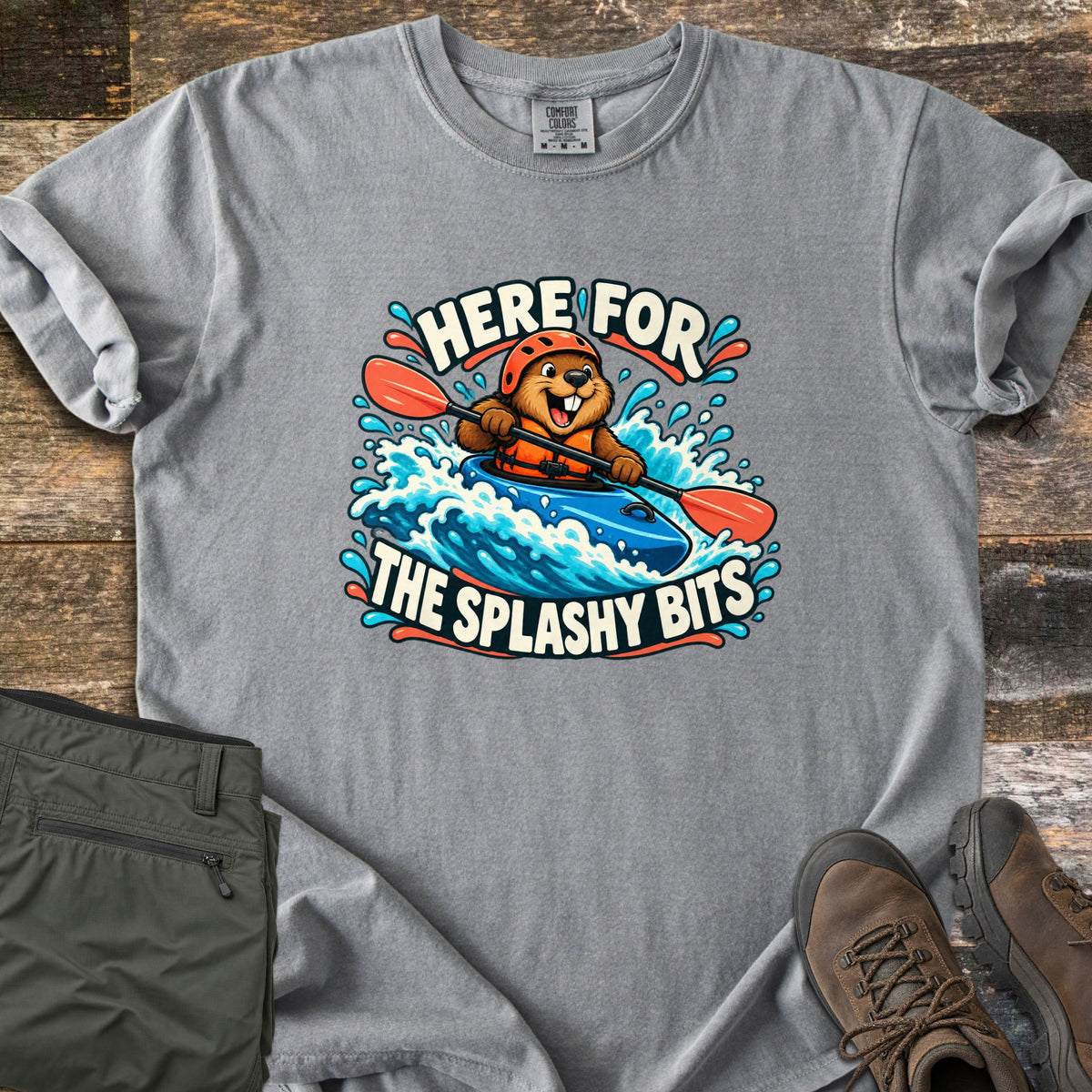 Splashy Bits Comfort Colors T-shirt in Grey, laid flat. A cartoon beaver in an orange helmet and life vest paddles a blue kayak through whitewater, framed by the words Here For The Splashy Bits. Olive pants and brown hiking boots lie beside it on weathered wood.