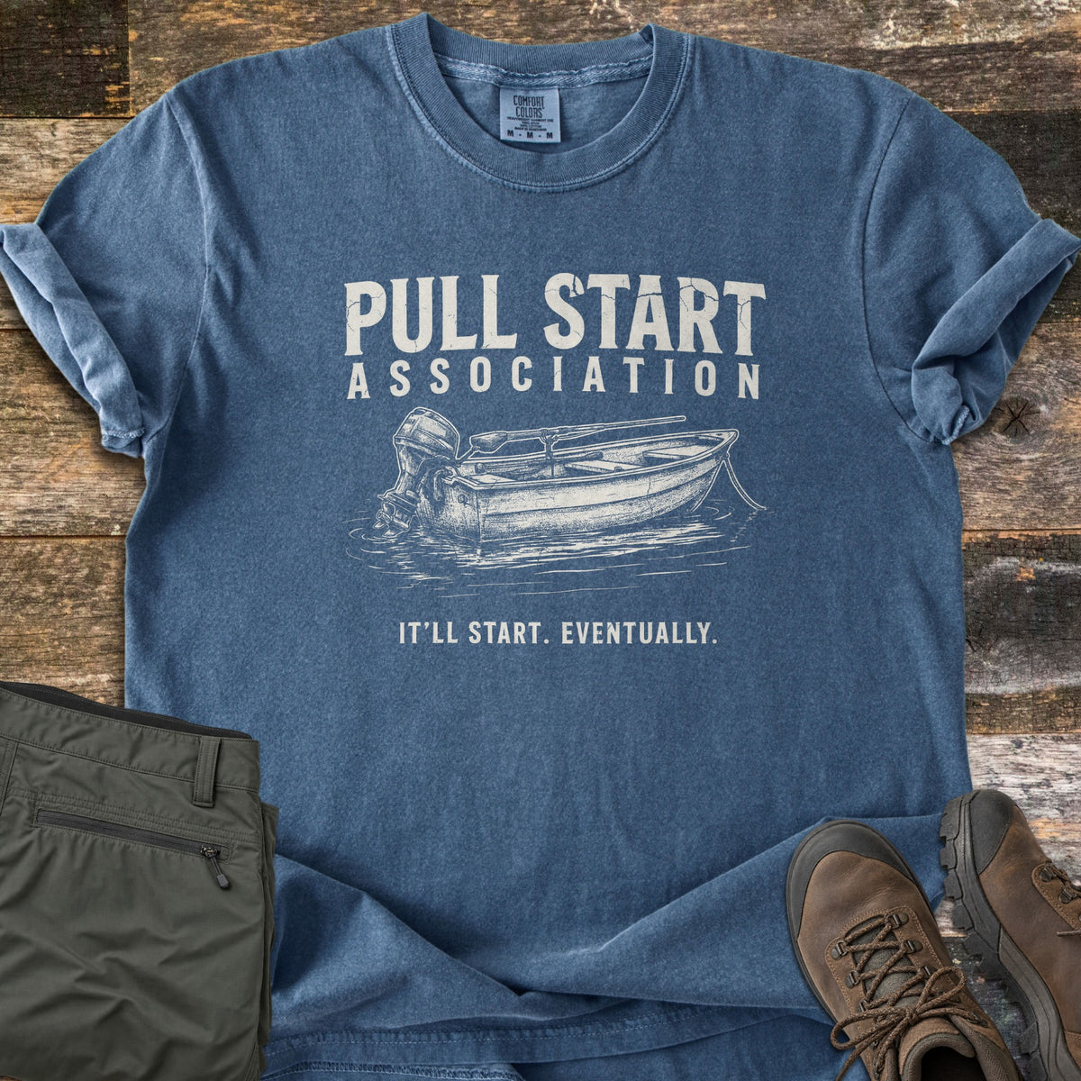Pull Start Association Comfort Colors T-shirt in Denim, laid flat. White distressed print of a small rowboat with an outboard motor on rippled water, under the words Pull Start Association and above It'll Start. Eventually. Olive pants and brown hiking boots lie beside it on weathered wood.