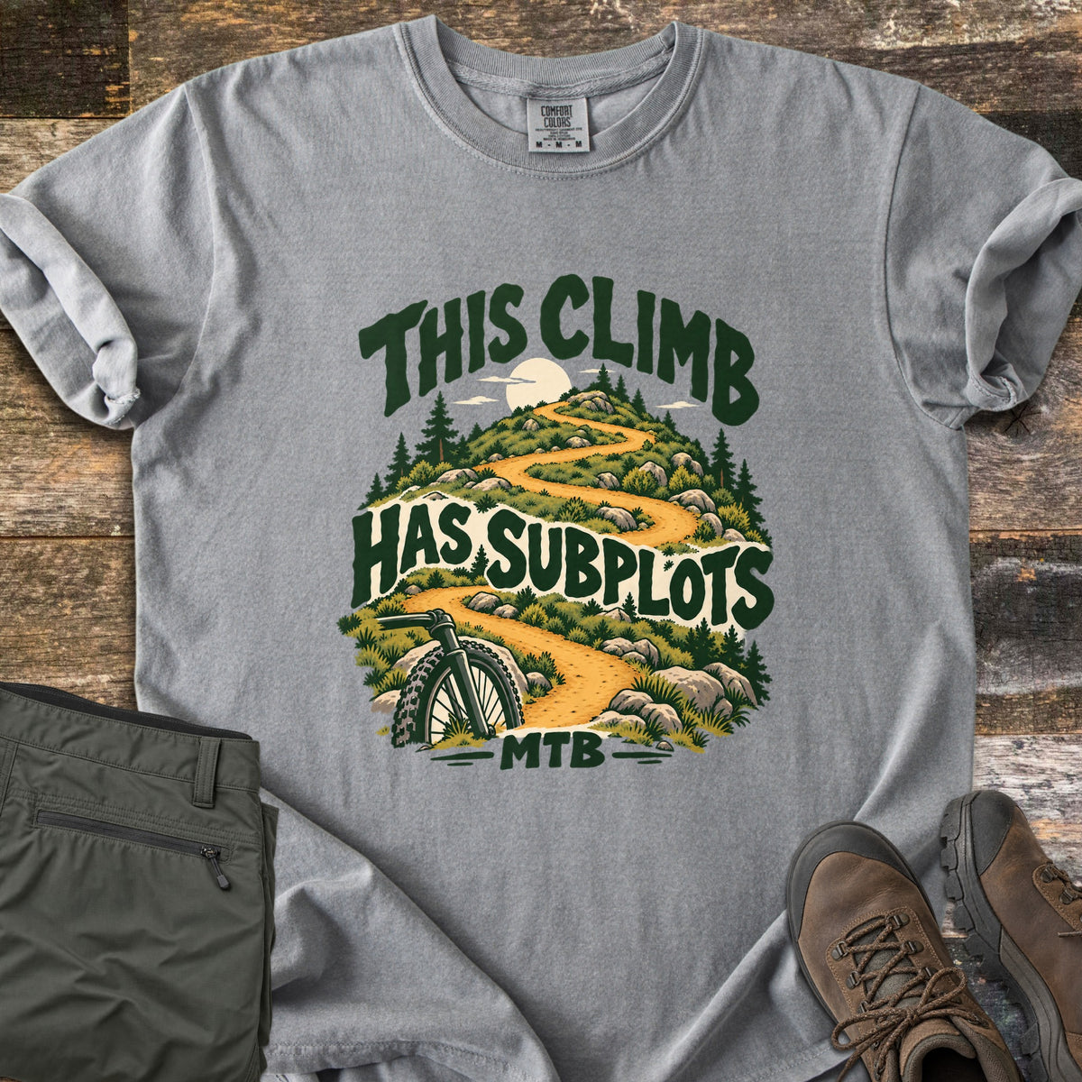 This Climb Has Subplots Comfort Colors T-shirt in Grey, laid flat. A winding dirt trail climbs a rocky hill with pine trees and a pale sun, a mountain bike wheel in front, under the words This Climb Has Subplots MTB. Olive pants and brown hiking boots lie on dark wood boards.