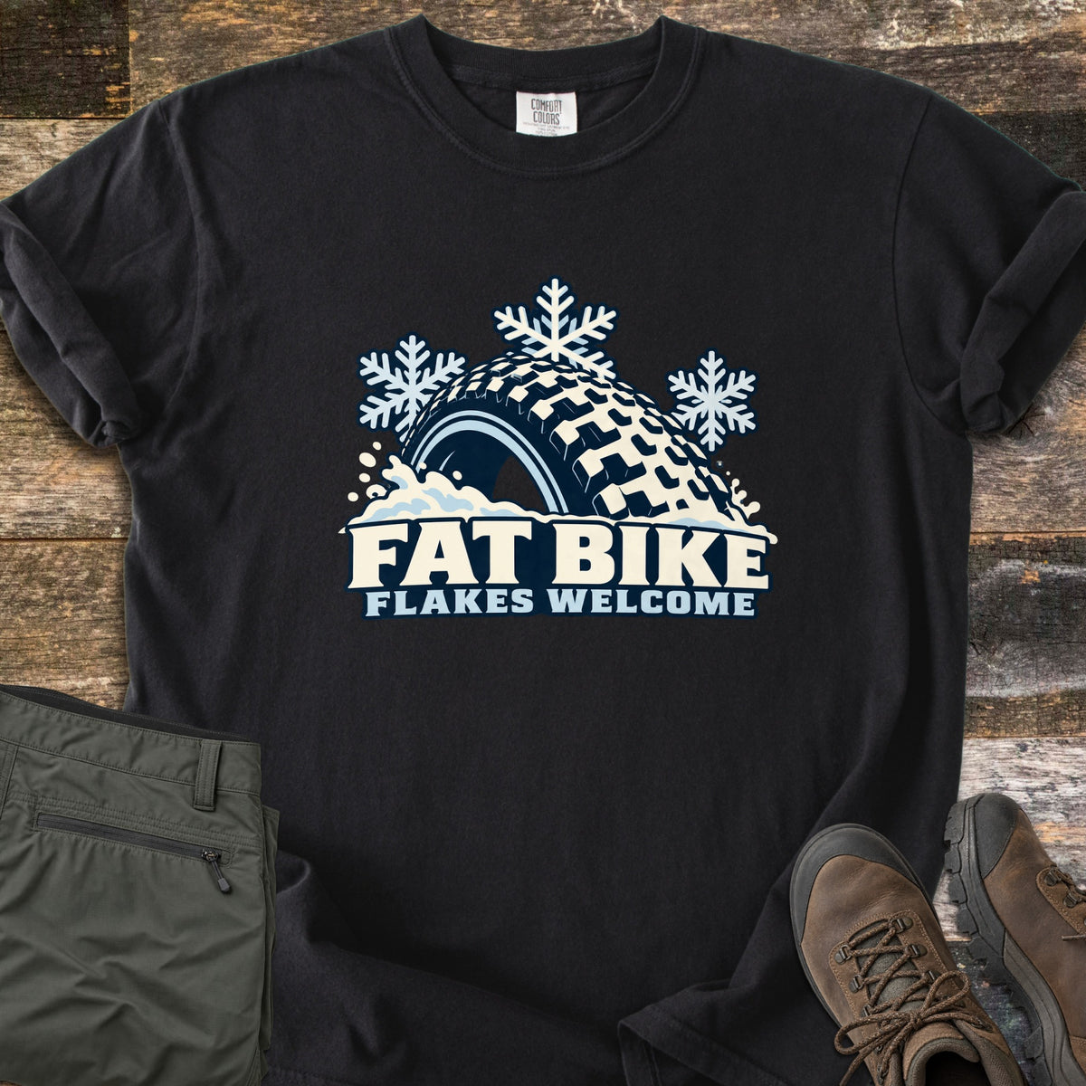 Fat Bike Flakes Welcome Comfort Colors T-shirt in Black, laid flat. A fat bike tire half buried in snow with three snowflakes above it, over the words FAT BIKE FLAKES WELCOME. Sleeves are rolled, on a wood floor with folded olive pants and brown hiking boots.