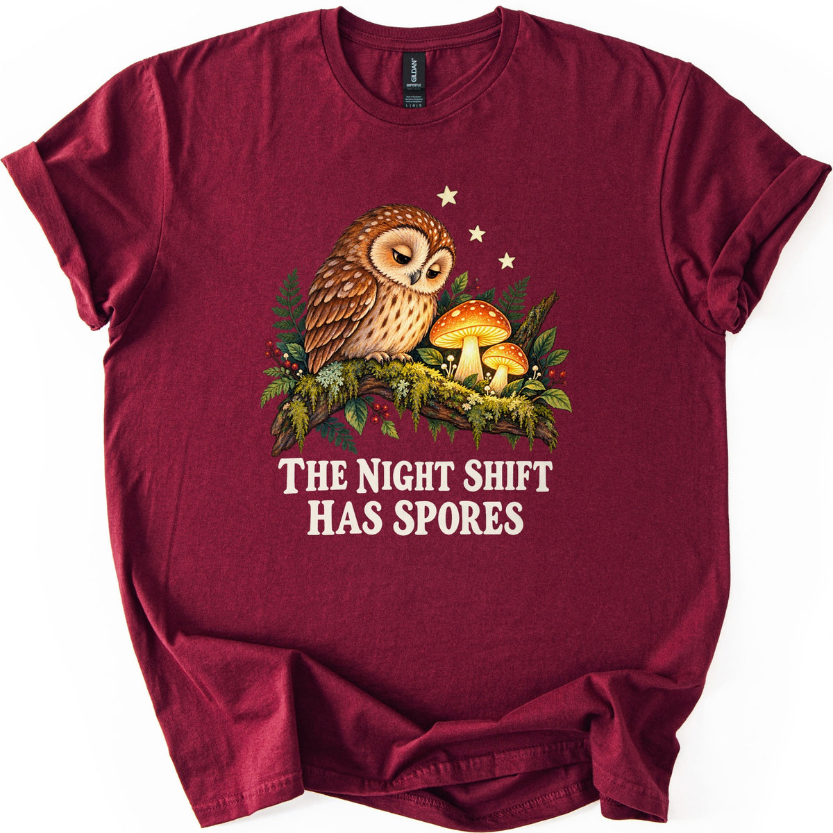 The Night Shift Has Spores Comfort Colors T-shirt in Maroon, laid flat. A sleepy brown owl perches on a mossy branch beside two glowing orange mushrooms, with ferns, red berries and small stars, above the words The Night Shift Has Spores in cream. Shown on white.