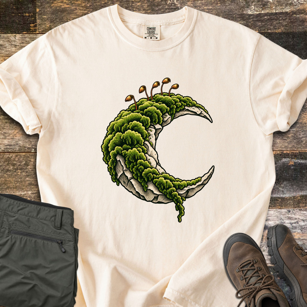 Moss Moon Comfort Colors T-shirt in Ivory, laid flat. A pale stone crescent moon blanketed in green moss, with thin brown spore stalks rising from the top. Sleeves rolled, on weathered wood boards beside folded olive hiking pants and brown hiking boots.
