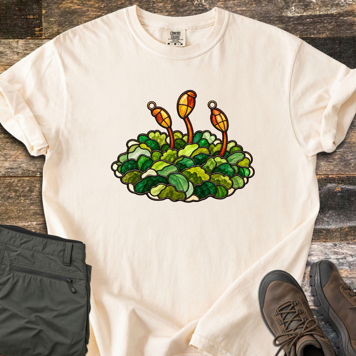Stained Glass Moss Comfort Colors T-shirt in Ivory, laid flat. A stained glass style moss mound in greens with three orange spore capsules on stalks above it, no text. Folded olive green pants and brown leather hiking boots rest beside it on weathered wood planks.