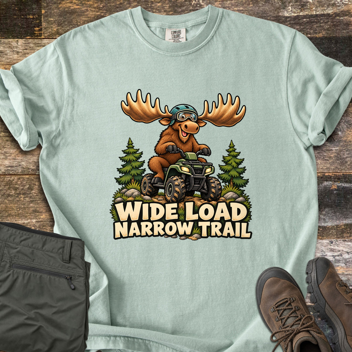 Wide Load Narrow Trail Comfort Colors T-shirt in Bay, laid flat. A smiling cartoon moose in a helmet and goggles rides a green ATV on a dirt trail flanked by pine trees above the words Wide Load Narrow Trail. Olive pants and brown hiking boots rest on weathered wood boards.