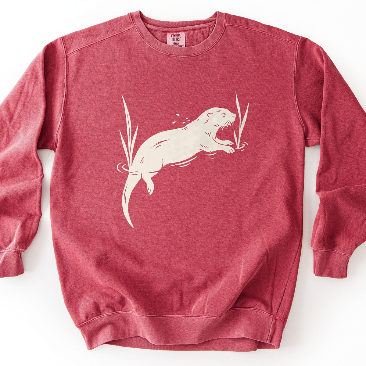 Swimming Otter Comfort Colors crewneck sweatshirt in Crimson, laid flat. A cream block-print style otter swims with its head raised, splashing droplets, between two tall reeds with ripples around them. No words are printed. The sweatshirt lies on a white background.