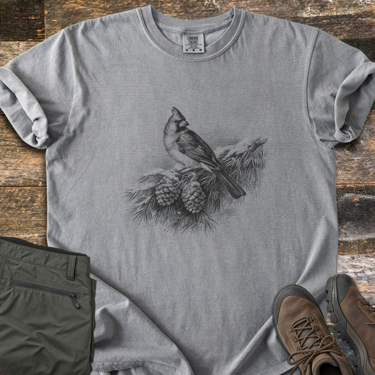 Cardinal On A Snowy Bough Comfort Colors T-shirt in Grey, laid flat. A grayscale drawing of a cardinal perched on a snow-covered pine branch with two pinecones, with no words. Sleeves rolled, on weathered wood planks beside olive hiking pants and brown hiking boots.