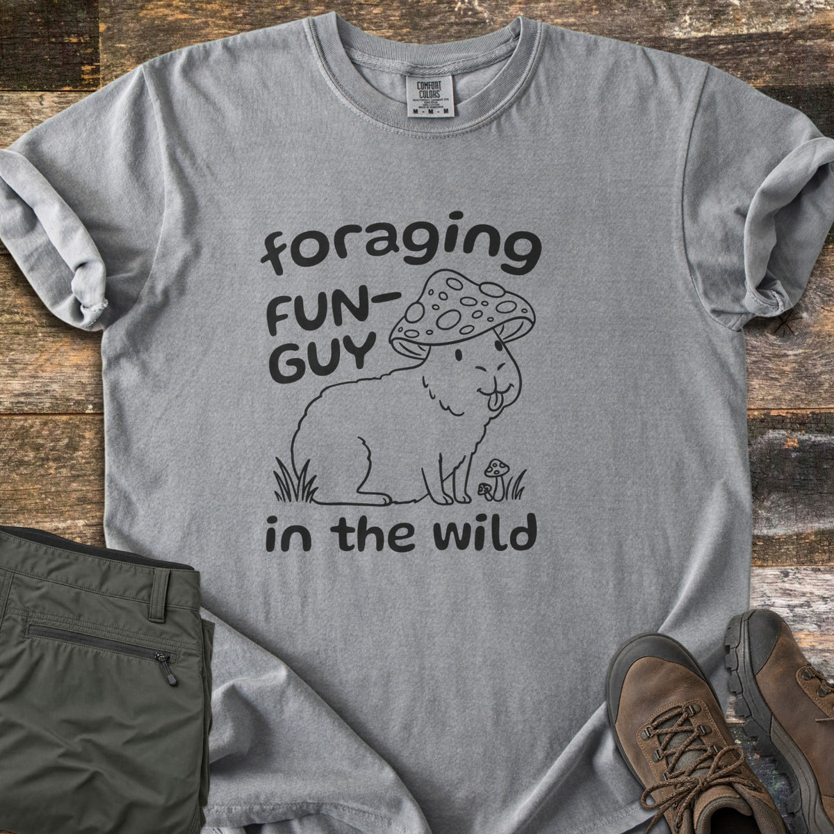 Foraging Fun-Guy in the Wild Comfort Colors T-shirt in Grey, laid flat. A black line drawing of a capybara wearing a spotted mushroom cap, tongue out, sits in grass by tiny mushrooms between the words foraging FUN-GUY in the wild. Olive hiking pants and brown boots lie on weathered wood.