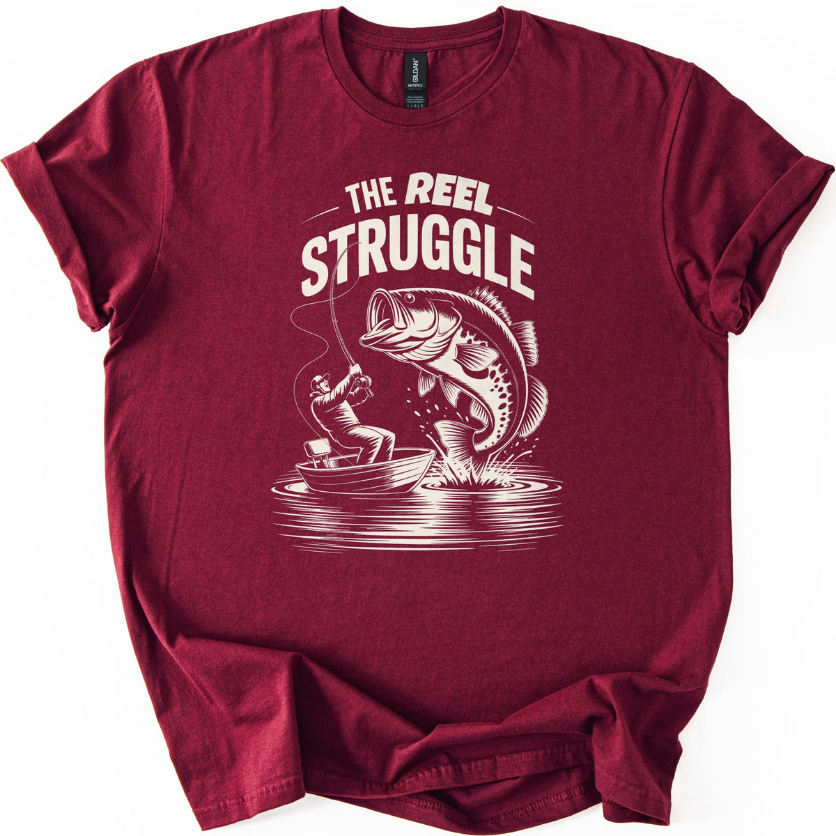 Reel Struggle Comfort Colors T-shirt in Maroon, laid flat. Cream print of the words The Reel Struggle above a fisherman in a small boat leaning back on a bent rod as a large bass leaps from the splashing water. The sleeves are rolled, on a white background.