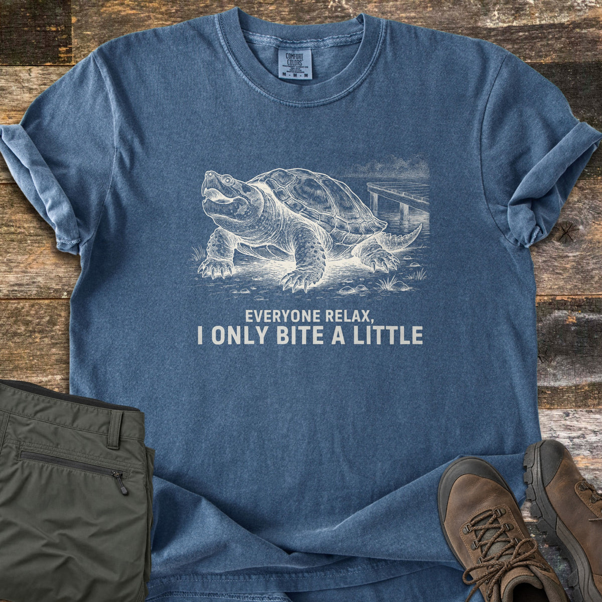 Everyone Relax I Only Bite A Little Comfort Colors T-shirt in Denim, laid flat. A white sketch of a snapping turtle with open mouth on a lakeshore by a dock, above the words Everyone Relax, I Only Bite A Little. Sleeves rolled, on wood planks with olive pants and brown hiking boots.