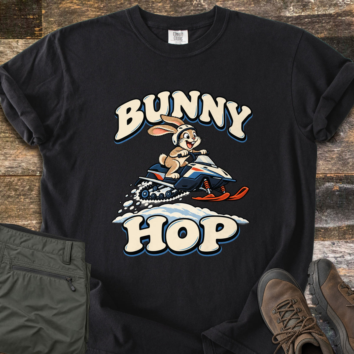 Bunny Hop Comfort Colors T-shirt in Black, laid flat. A cartoon bunny in a white helmet rides a navy, white and orange snowmobile over a snowbank between the words Bunny Hop. Folded green pants and brown hiking boots lie beside it on a wood floor.