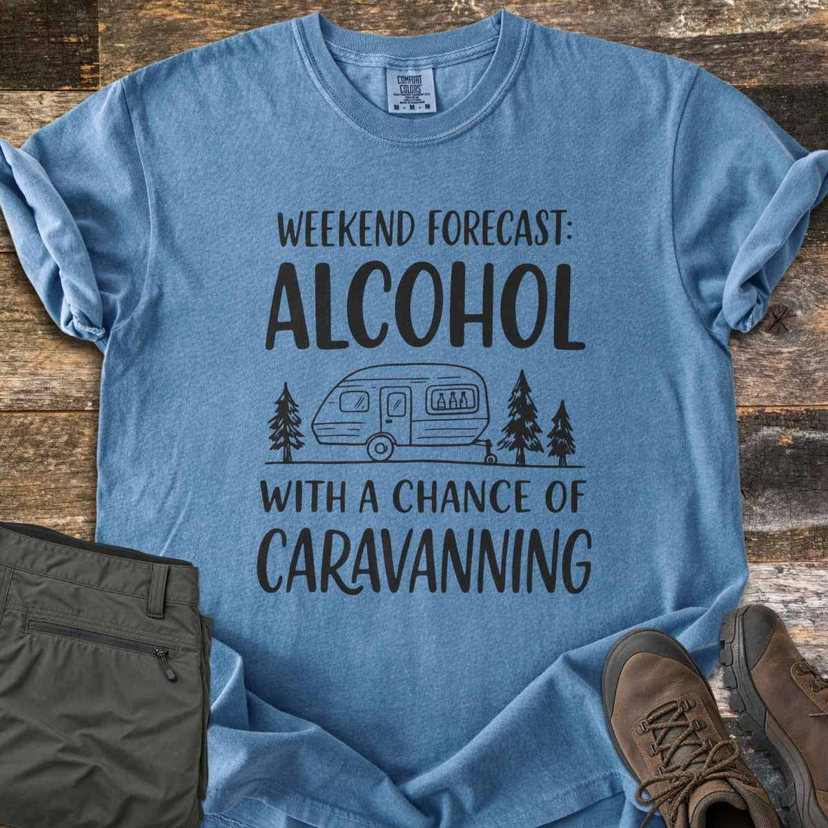 Alcohol With a Chance of Caravanning Comfort Colors T-shirt in Blue Jean, laid flat. Black print reads Weekend Forecast: Alcohol above a caravan with bottles in its window between pine trees, then With a Chance of Caravanning. Olive trousers and brown hiking boots sit on dark wood planks.