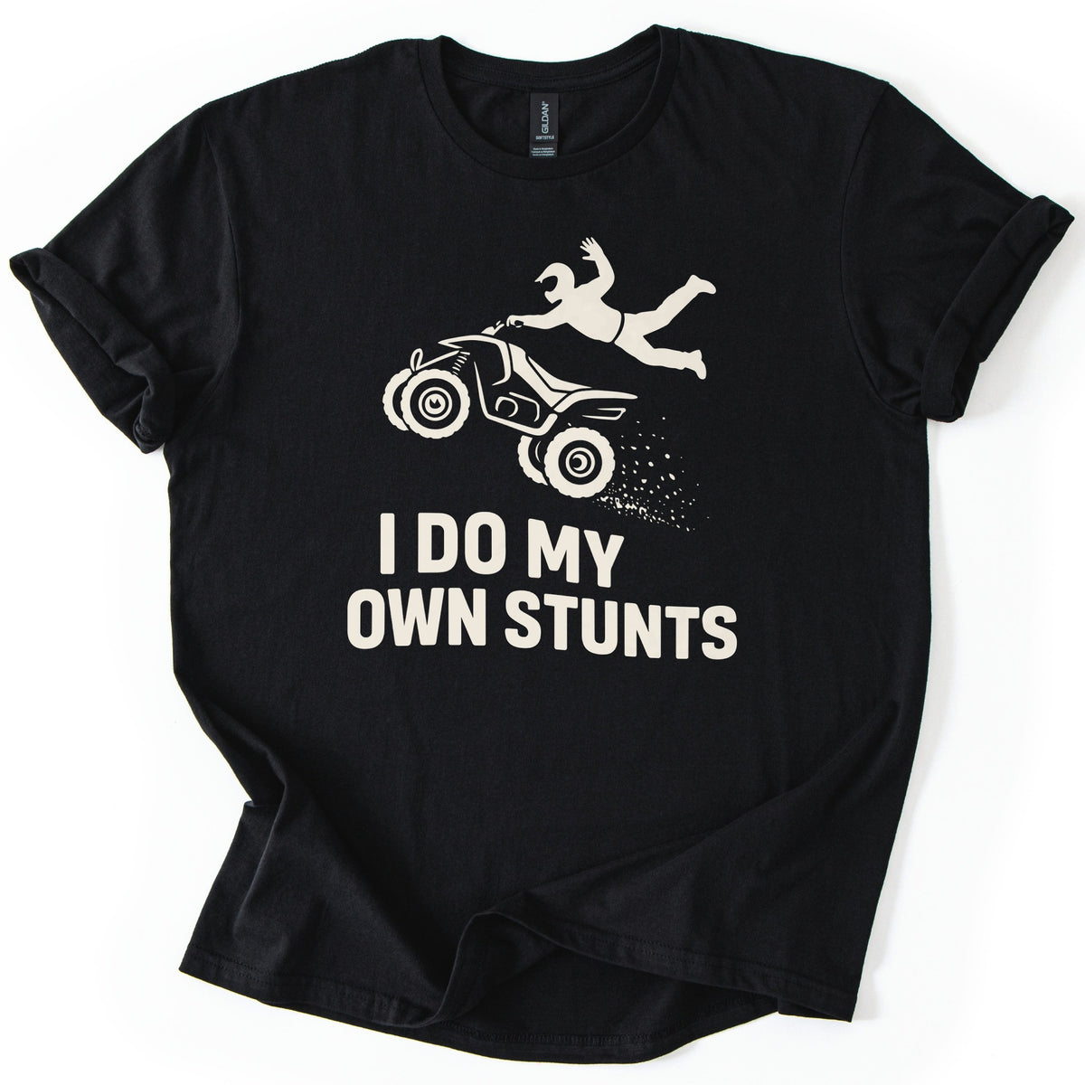 ATV Stunts Comfort Colors T-shirt in Black, laid flat. A cream silhouette of a helmeted rider flying off the back of an ATV as it kicks up dirt, above the words I DO MY OWN STUNTS. The shirt lies on a white background with its sleeves cuffed.