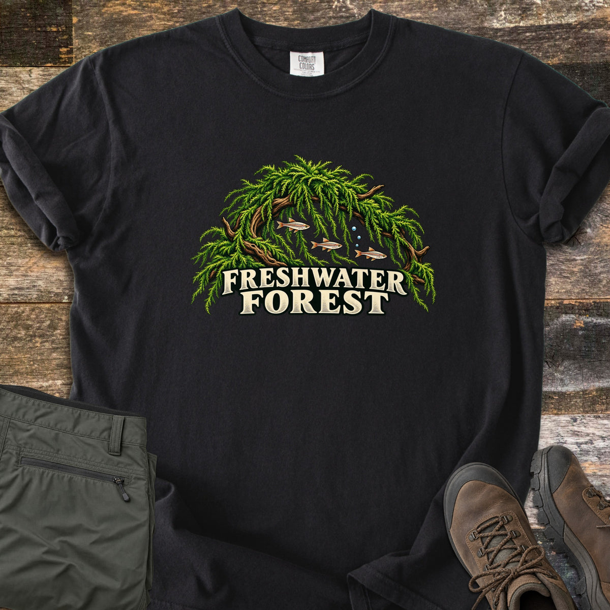 Freshwater Forest Comfort Colors T-shirt in Black, laid flat. Three small fish swim under a driftwood arch covered in green moss, above the cream words Freshwater Forest. Folded olive pants and brown hiking boots lie beside the shirt on worn wood boards.