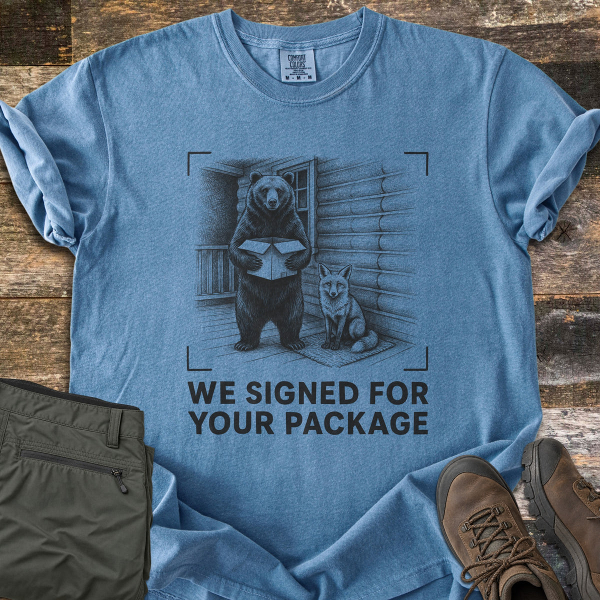 We Signed For Your Package Comfort Colors T-shirt in Blue Jean, laid flat. A black-ink sketch shows a bear holding an open box beside a seated fox on a log cabin porch, framed by camera corner marks above the words We Signed For Your Package. Olive pants and brown hiking boots sit on wood planks.