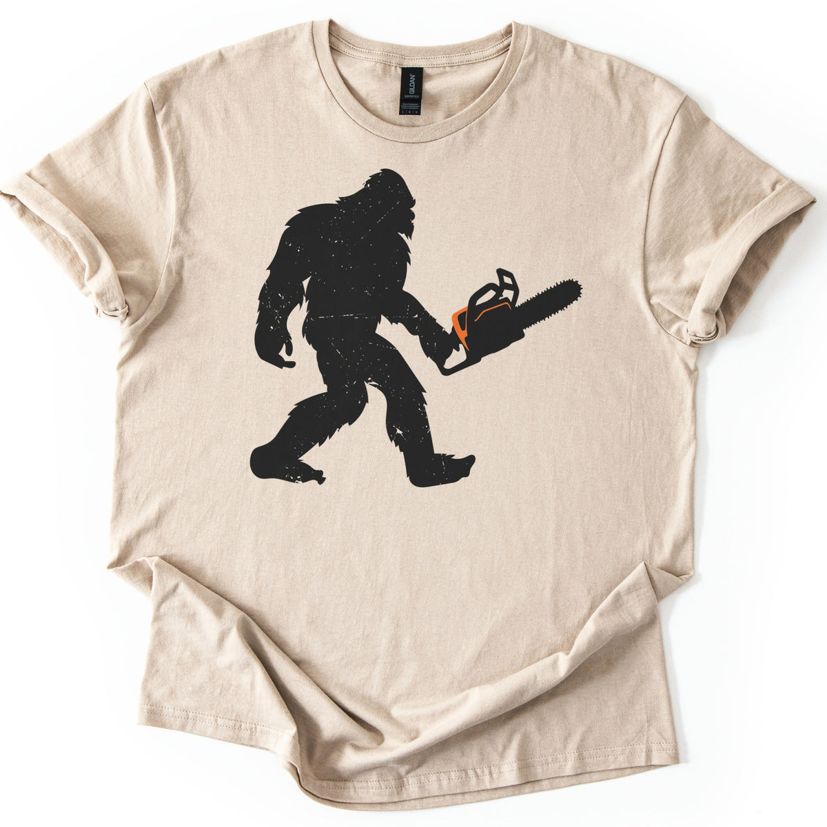 Lumber Squatch Comfort Colors T-shirt in Sand, laid flat. A distressed black silhouette of Bigfoot walking mid-stride while holding a chainsaw with an orange accent, with no words printed. The shirt has cuffed sleeves and lies on a white background.