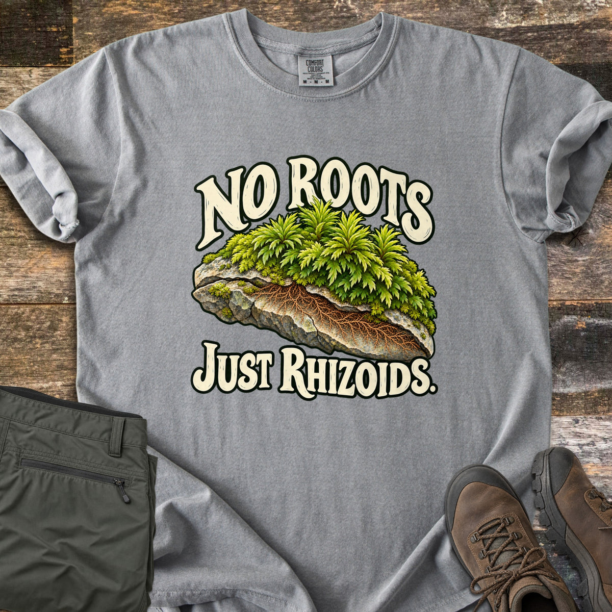 No Roots Just Rhizoids Comfort Colors T-shirt in Grey, laid flat. Green moss sprouts from a cracked rock with brown rhizoids threading underneath, between the words No Roots Just Rhizoids. On rustic wood beside folded olive hiking pants and brown hiking boots.