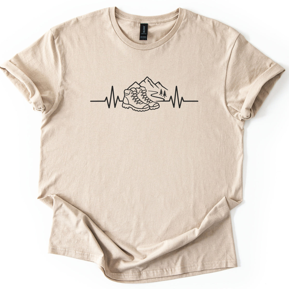 Hiking Heartbeat Comfort Colors T-shirt in Sand, laid flat. A black line drawing of a heartbeat line that turns into a pair of hiking boots in front of mountains, a winding trail and two pine trees; no words. Sleeves are cuffed on a white background.