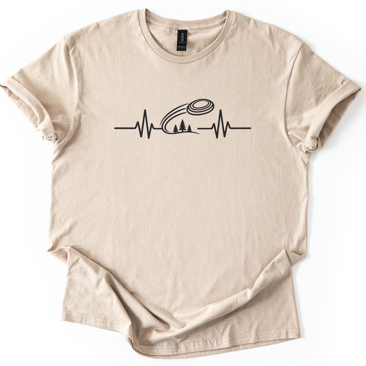 Trap Shooting Heartbeat Comfort Colors T-shirt in Sand, laid flat. A black heartbeat line with a clay target flying above three pine trees on a hill, with no words. The shirt lies on a white background with its sleeves rolled and the hem loosely folded.