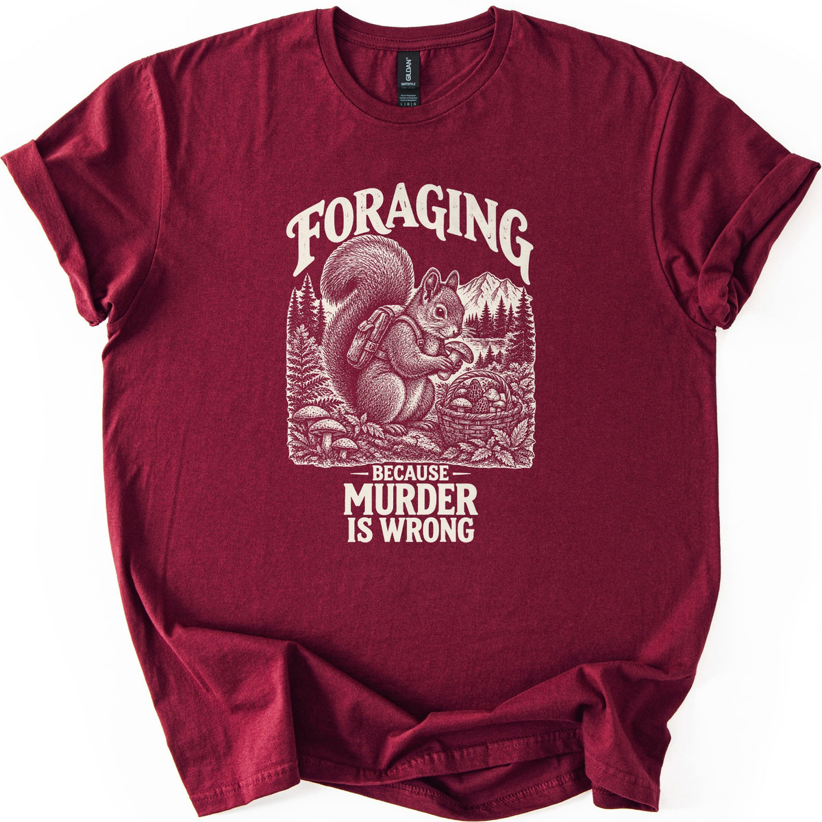 Foraging Murder Squirrel Comfort Colors T-shirt in Maroon, laid flat. Foraging arches over a cream etched squirrel wearing a backpack and holding a mushroom beside a basket of mushrooms, with pines and a mountain behind. Below: Because Murder Is Wrong. Cuffed sleeves, white background.