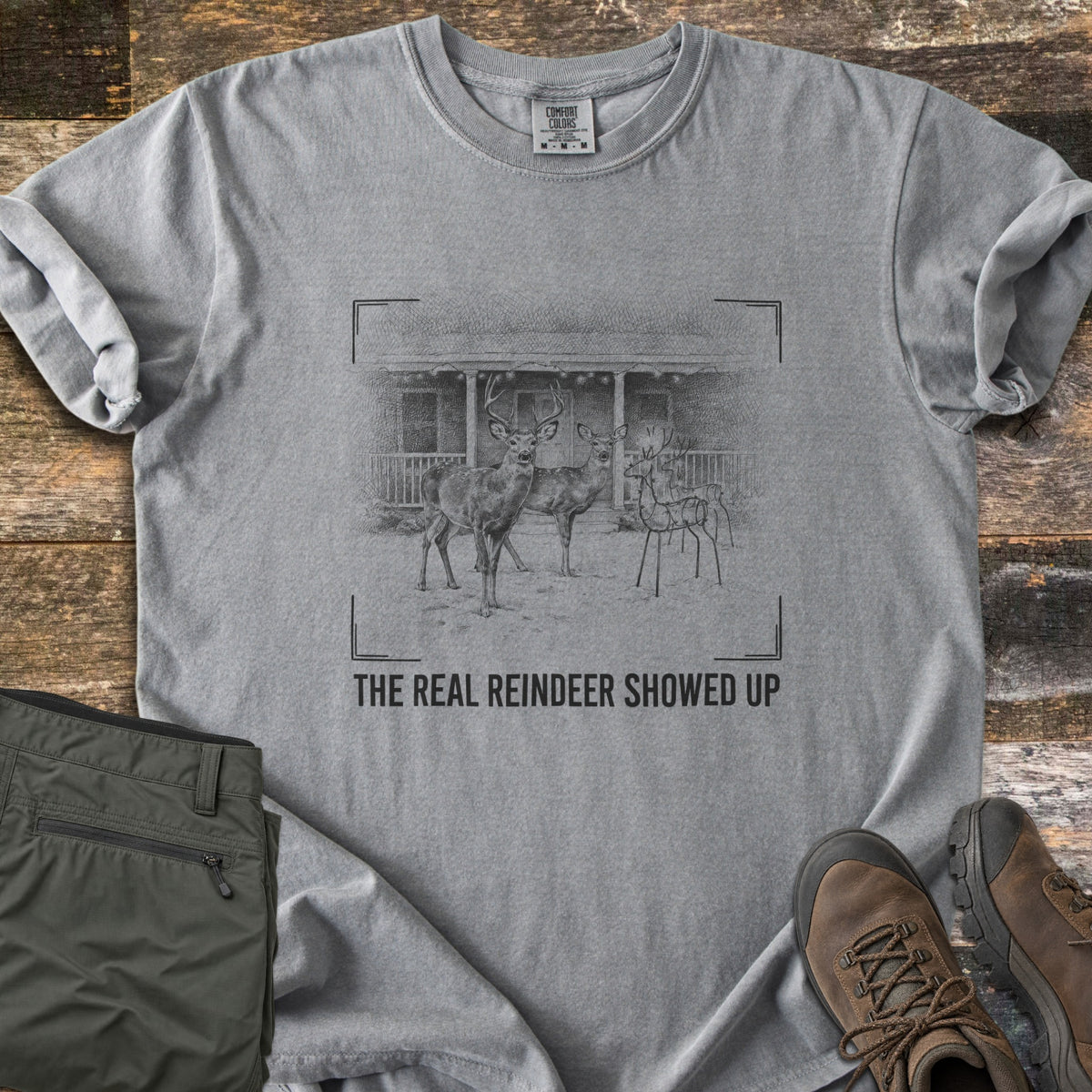 The Real Reindeer Showed Up Comfort Colors T-shirt in Grey, laid flat. Viewfinder corners frame a sketch of two real deer and a wire reindeer decoration in a snowy yard by a porch with string lights, above the words The Real Reindeer Showed Up. Folded olive pants and hiking boots lie beside it.