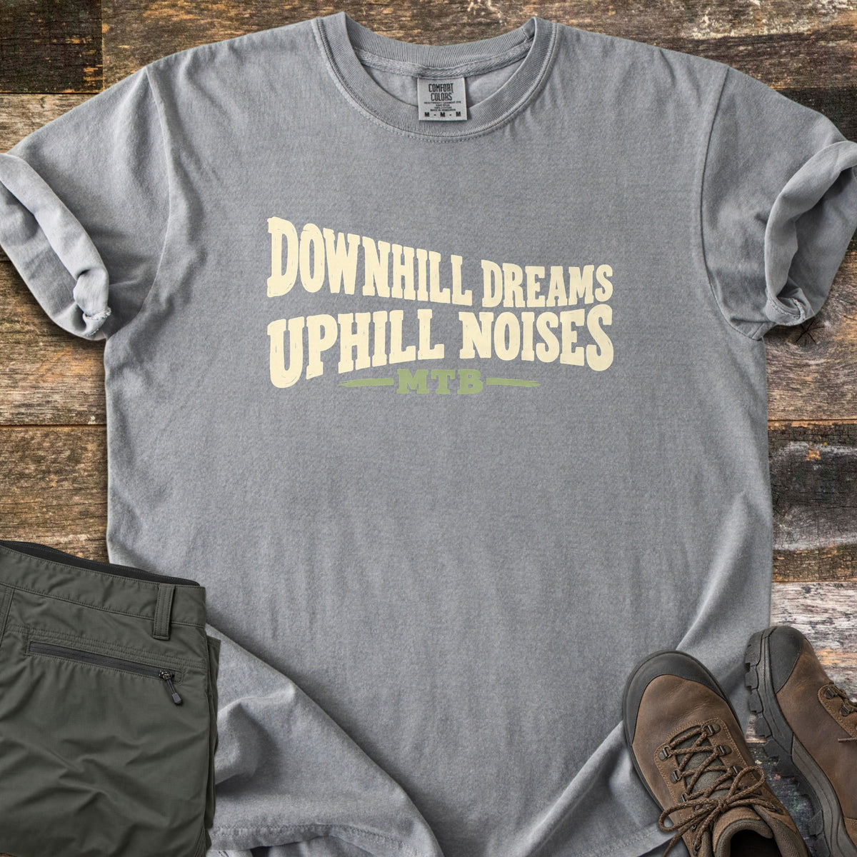 Downhill Dreams Uphill Noises Comfort Colors T-shirt in Grey, laid flat. Cream wavy block letters read Downhill Dreams Uphill Noises above MTB in green between two green lines. Sleeves rolled on rustic wood planks, next to folded olive hiking pants and brown hiking boots.