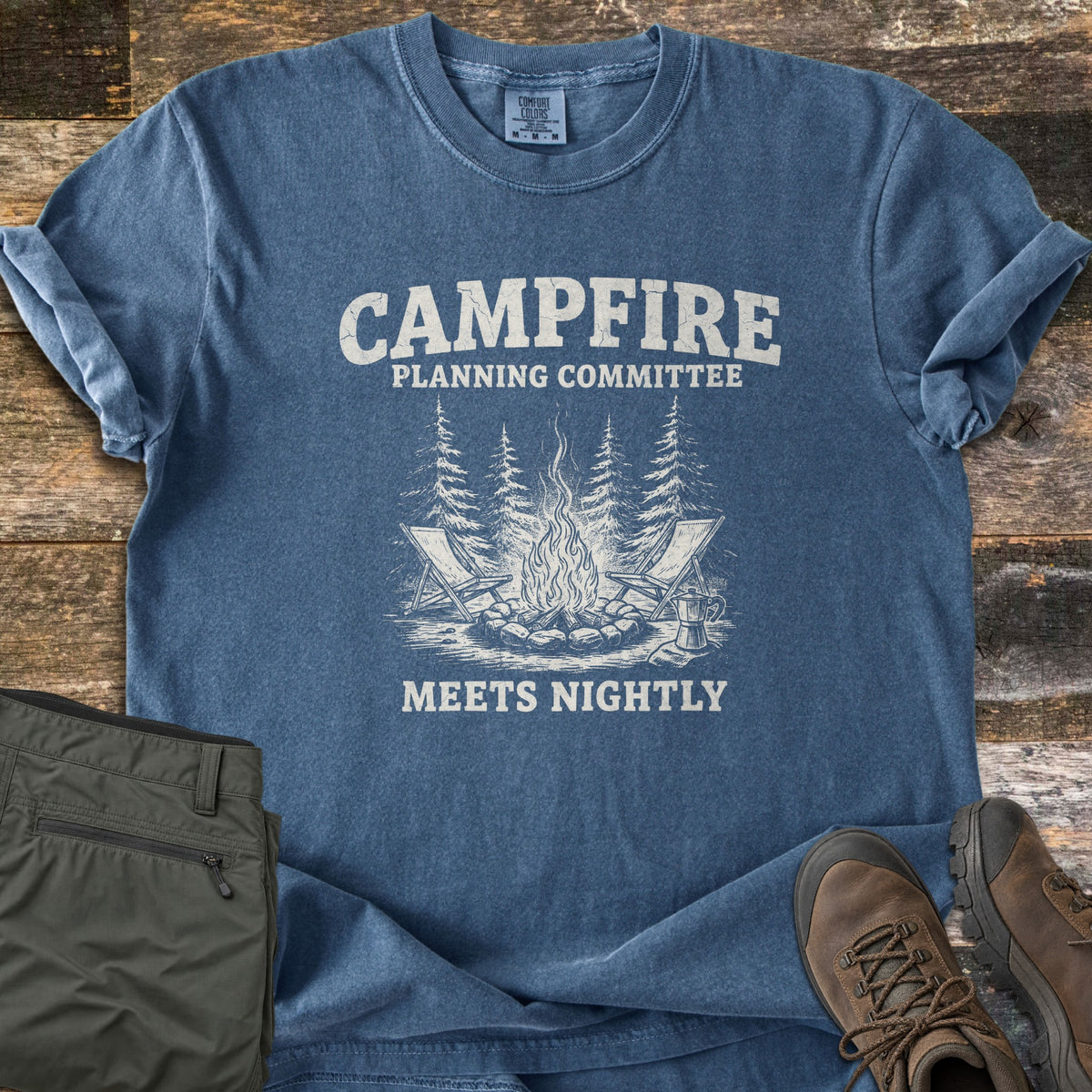 Campfire Planning Committee Comfort Colors T-shirt in Denim, laid flat. White distressed print: CAMPFIRE, PLANNING COMMITTEE above two camp chairs, a campfire, pine trees and a coffee pot, then MEETS NIGHTLY. Olive pants and brown hiking boots sit on weathered wood planks.
