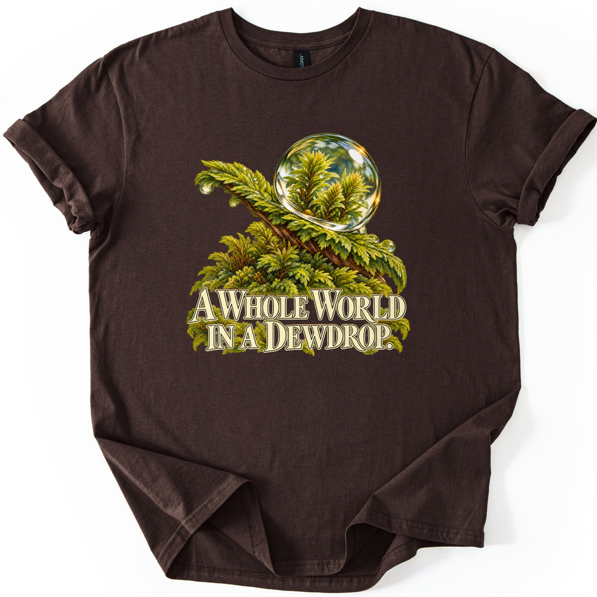 A Whole World in a Dewdrop Comfort Colors T-shirt in Dark Chocolate, laid flat. A shiny dewdrop perches on a lush green moss branch with small droplets along it, above cream lettering that reads A Whole World in a Dewdrop. Shown on a white background with sleeves rolled.