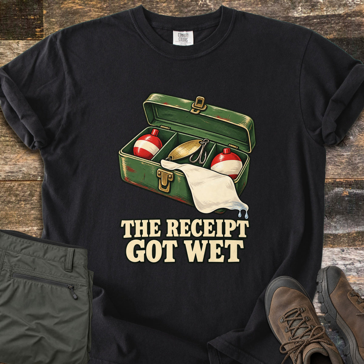 The Receipt Got Wet Comfort Colors T-shirt in Black, laid flat. An open green tackle box holds red and white bobbers and a gold spoon lure, with a dripping white paper hanging out, above the words The Receipt Got Wet. Olive pants and brown hiking boots sit on weathered wood.