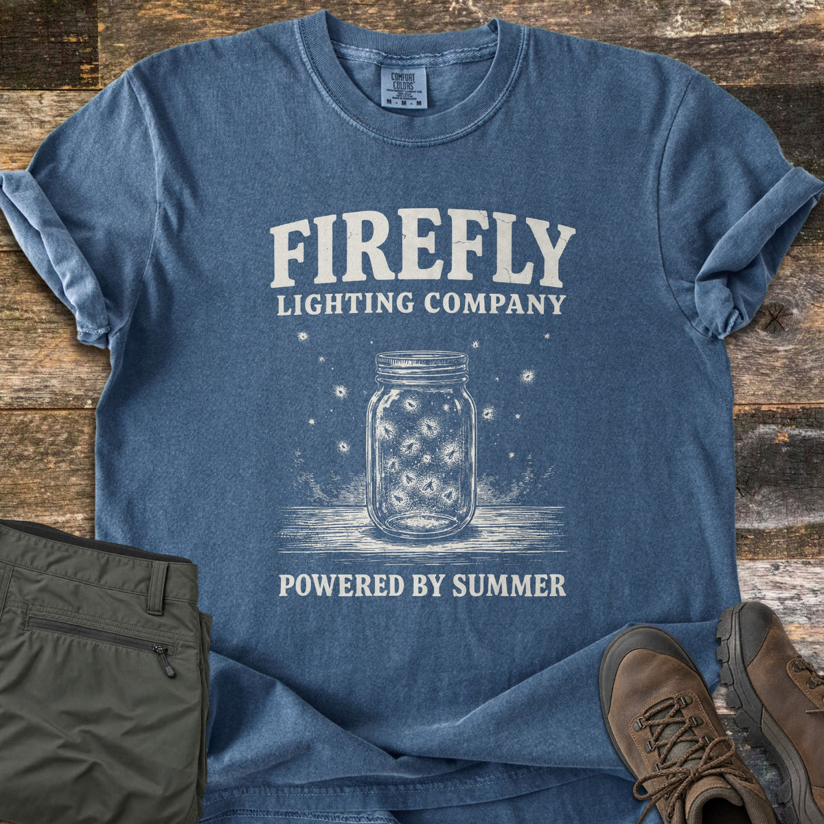 Firefly Lighting Company Comfort Colors T-shirt in Denim, laid flat. A sketched mason jar of glowing fireflies sits under the words Firefly Lighting Company and above Powered By Summer. Olive cargo pants and brown hiking boots lie beside it on weathered wood planks.