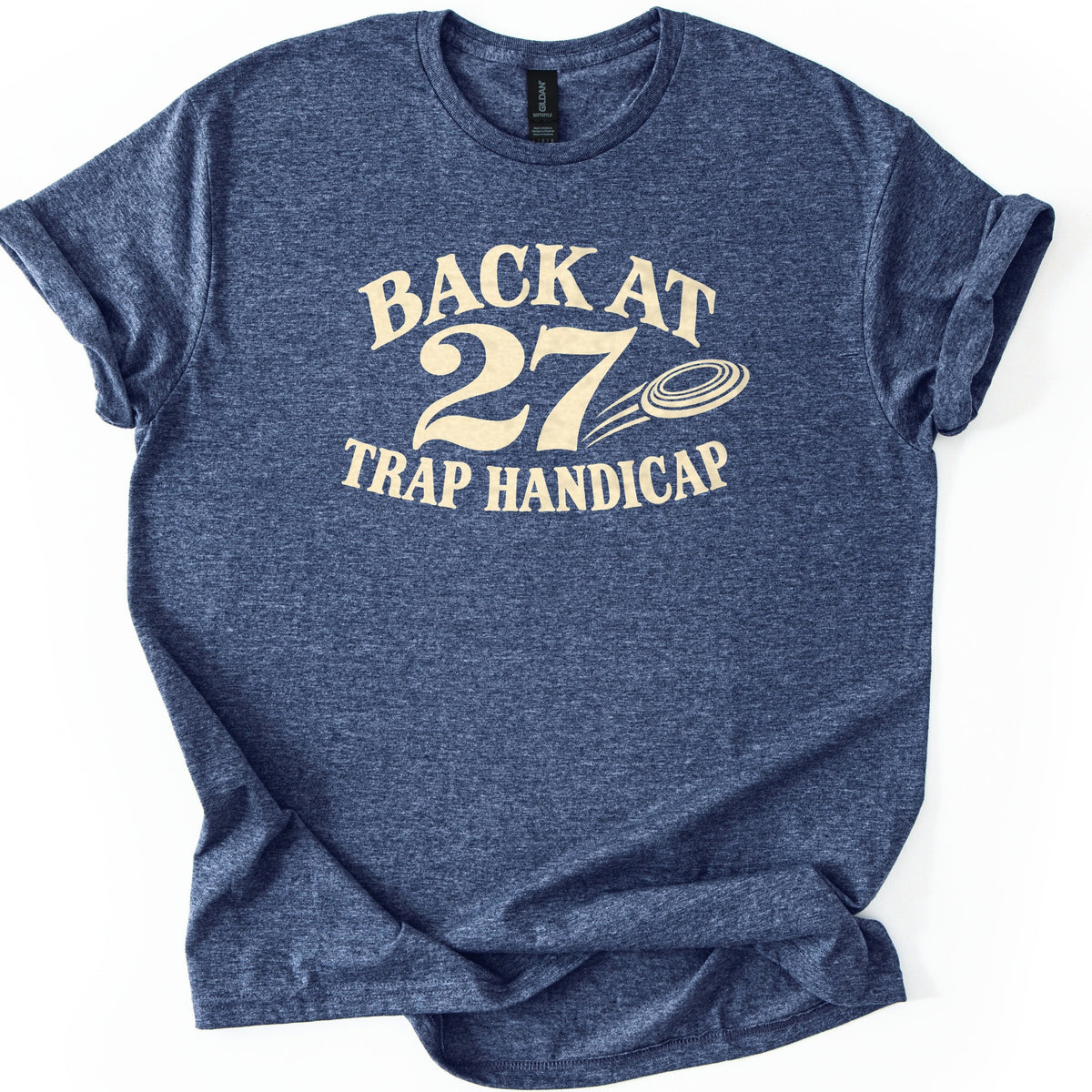 Back at 27 Trap Handicap Comfort Colors T-shirt in Heather Navy, laid flat. Cream lettering reads Back at 27 Trap Handicap, with a flying clay target and motion lines next to the large 27. The shirt has rolled sleeves and lies on a white background with the hem folded at one side.