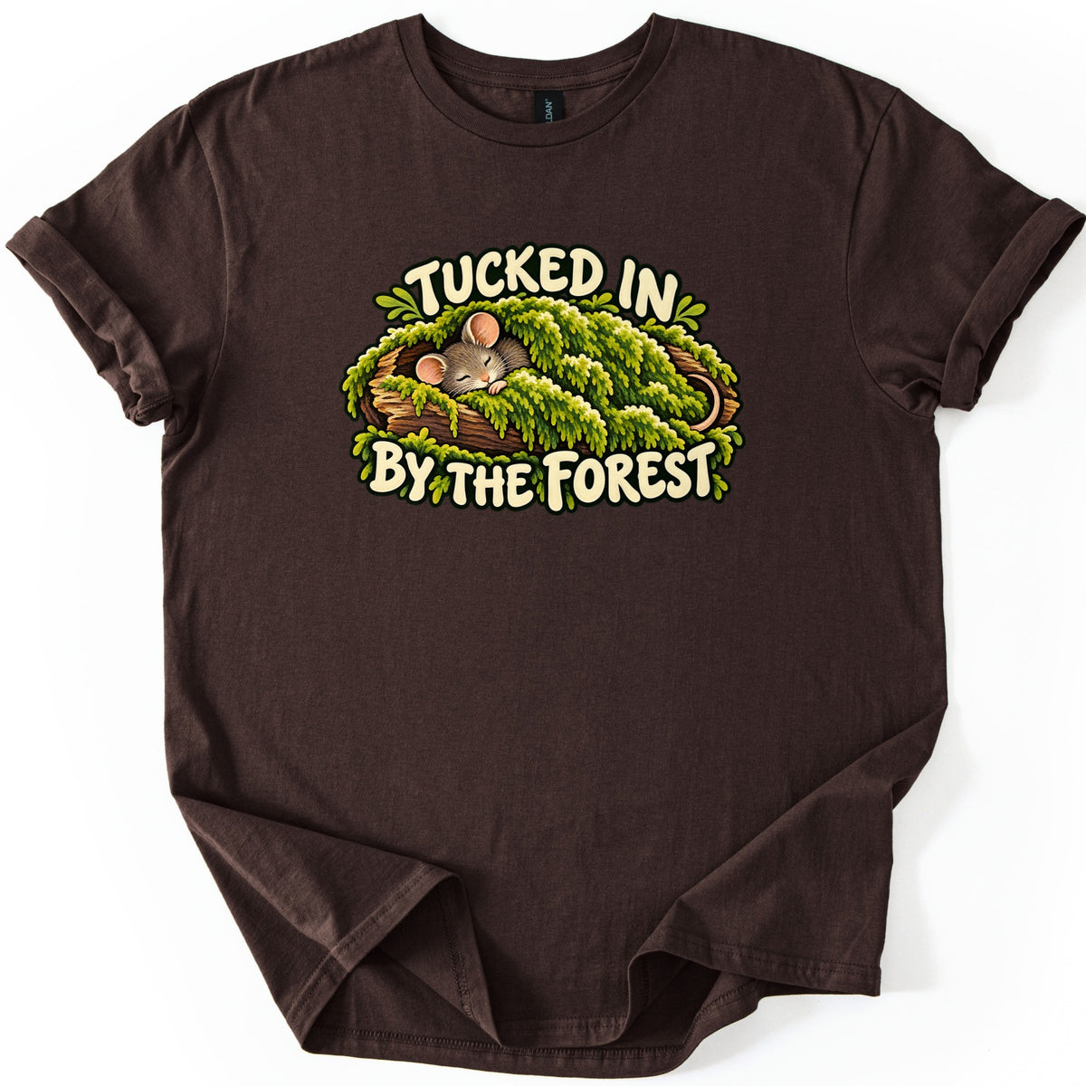 Tucked in by the Forest Comfort Colors T-shirt in Dark Chocolate, laid flat. A sleeping gray mouse with pink ears lies under a blanket of green moss in a hollow log, between cream words reading Tucked in By the Forest. The shirt has rolled sleeves on a white background.