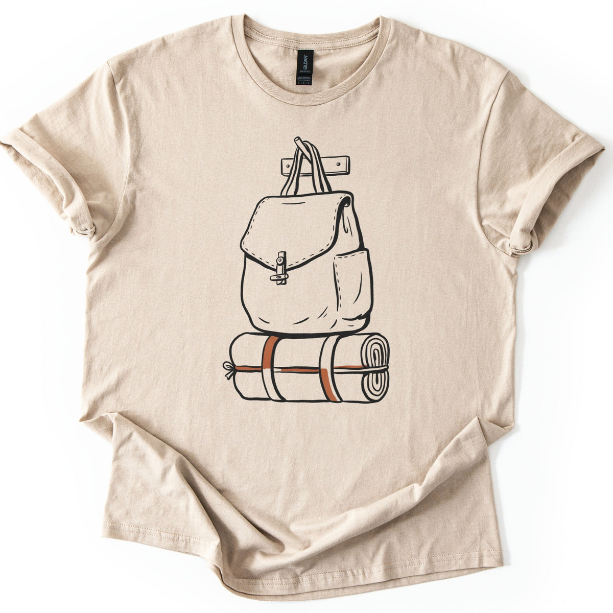 Backpack And Bedroll Comfort Colors T-shirt in Sand, laid flat. A black line drawing of a canvas backpack with a toggle-clasp flap hangs by its handles from a wall peg above a rolled bedroll tied with rust-red straps. No words. Cuffed sleeves on a white surface.